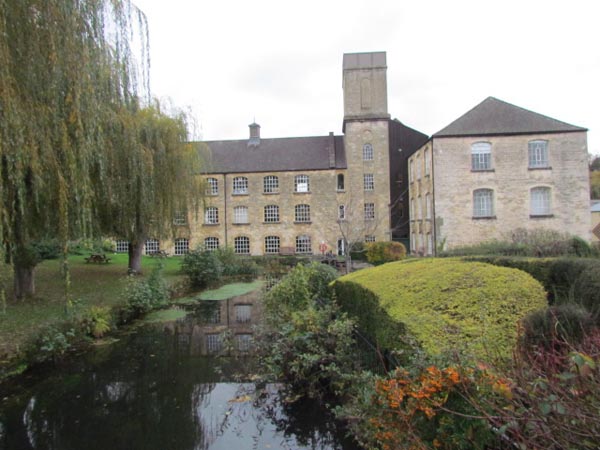 A three storey stone building, with a tower and additional building to one side can be seen with its mill pond in the foreground.