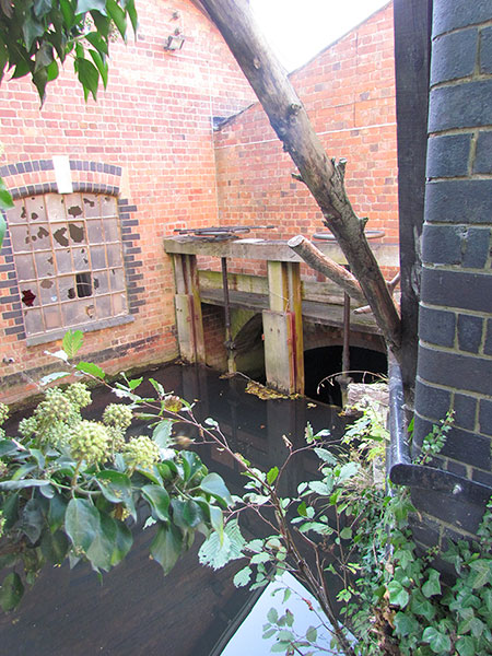 The view through a gap in the wall shows water pouring over a sluice and down to the River Frome beyond. Alongside is the broken old metal window to a former industrial building.
