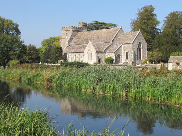 A stone church with a low tower and battlements sits next to the canal. There are tall water plants – irises and bulrushes growing next to the water.